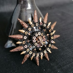 Könnte beinhalten: Eine schwarze und goldene Perlen-Sonnenstrahlen-Brosche mit einem Totenkopf in der Mitte. Die Brosche hat ein stacheliges, strukturiertes Design.