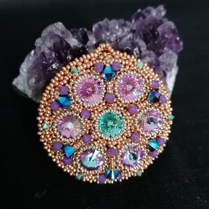 May include: A copper-toned beaded pendant with a circular design featuring a variety of iridescent crystals in shades of pink, purple, blue, and green.