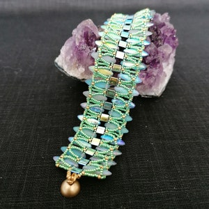 Könnte beinhalten: Ein grünes und schillerndes Perlenarmband mit einer goldenen Perle am Ende. Das Armband liegt auf einem violetten Amethystkristall.