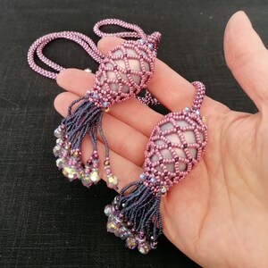 以下が含まれることがあります： ピンクとパープルの虹色のガラスビーズで作られたビーズネックレス2本。各ネックレスには、大きなビーズでできたケージのようなペンダントとタッセルが付いています。