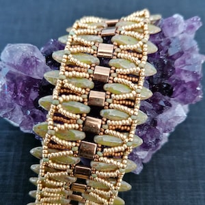 Könnte beinhalten: Ein Armband mit olivgrünen und goldenen Rocailles und rechteckigen goldfarbenen Metallakzenten. Das Armband wird auf einem lila Kristall präsentiert, was die detaillierte Perlenarbeit und den Farbkontrast hervorhebt.