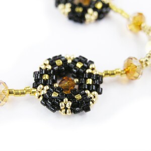Puede incluir: Primer plano de un collar de cuentas negras y doradas con cuentas de cristal color &aacute;mbar. El collar presenta un patr&oacute;n repetitivo de racimos de cuentas circulares.