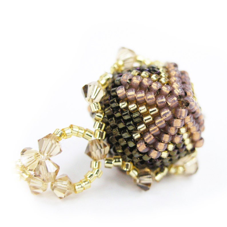 Puede incluir: Un amuleto de cuentas con un dise&ntilde;o geom&eacute;trico marr&oacute;n, dorado y morado. El amuleto est&aacute; unido a un anillo de cristales transparentes.