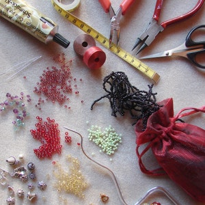 May include: A variety of beads, including red, black, green, and clear, are spread out on a white surface. There are also two pairs of pliers, a spool of thread, a tube of glue, and a small red pouch.
