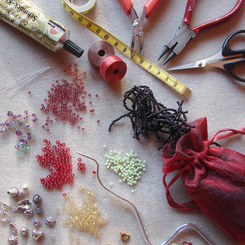 May include: A variety of beads, including red, black, green, and clear, are spread out on a white surface. There are also two pairs of pliers, a spool of thread, a tube of glue, and a small red pouch.