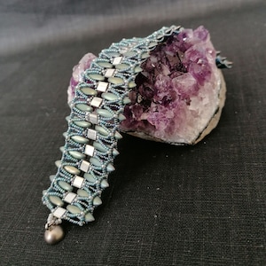 Könnte beinhalten: Ein Armband aus türkis- und silberfarbenen Perlen mit einem detaillierten, strukturierten Design. Das Armband hat einen kleinen Silberanhänger und wird auf einem lila Amethystkristall präsentiert. Der Hintergrund ist dunkel.