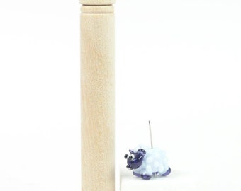 May include: A small glass sheep bead with a white body and blue accents. The bead is on a pin and is next to a wooden spool.