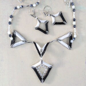 Puede incluir: Un collar de cuentas blanco y negro con un colgante geom&eacute;trico y pendientes a juego. El collar presenta una cadena larga con cuentas blancas y negras y un colgante triangular grande con un colgante m&aacute;s peque&ntilde;o en forma de diamante que cuelga de &eacute;l. Los pendientes son peque&ntilde;os y presentan un dise&ntilde;o geom&eacute;trico.