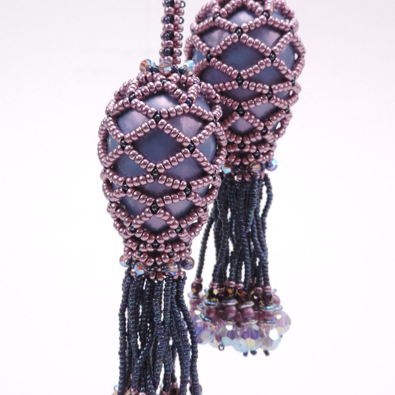 May include: A pair of beaded earrings in a purple and black colour scheme. The earrings feature a round, beaded ball with a lattice design and a tassel.