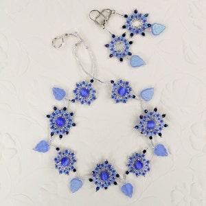Puede incluir: Un collar y pendientes de plata con cuentas de vidrio azul y morado y hojas plateadas. El collar tiene un dise&ntilde;o circular con una cuenta azul central y est&aacute; acentuado con hojas plateadas.