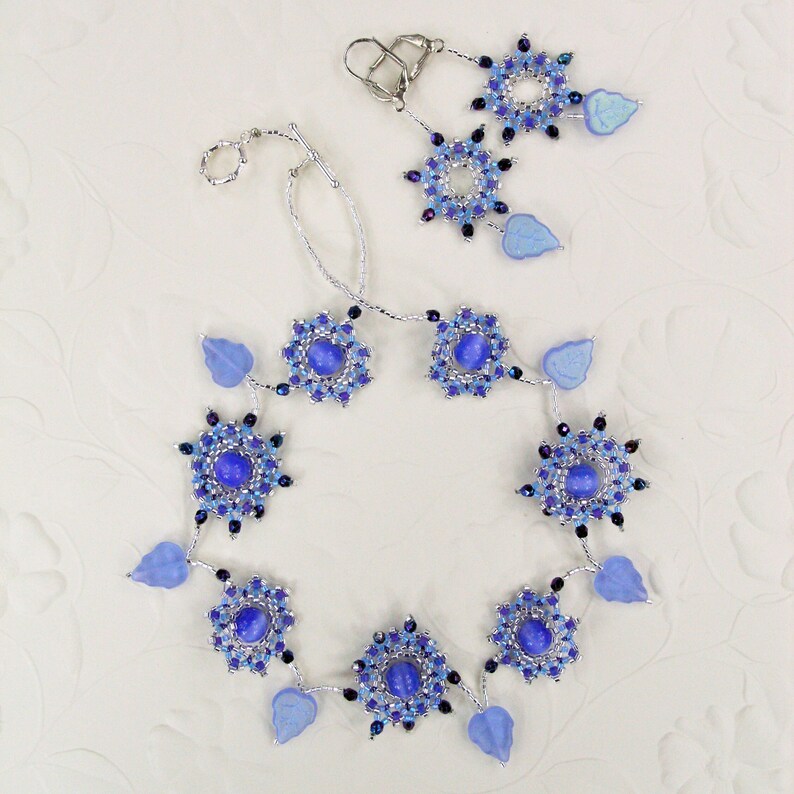 Puede incluir: Un collar y pendientes de plata con cuentas de vidrio azul y morado y hojas plateadas. El collar tiene un dise&ntilde;o circular con una cuenta azul central y est&aacute; acentuado con hojas plateadas.