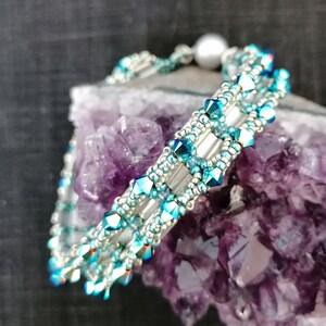 Könnte beinhalten: Ein silbernes und türkisfarbenes Armband mit Perlenakzent. Das Armband ist aus kleinen, facettierten Perlen gefertigt und wird auf einem violetten Amethystkristall präsentiert.