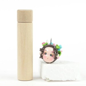 Könnte beinhalten: Ein kleiner, handgemachter Polymer Clay Puppenkopf mit braunen Haaren, blauen Augen und einer rosa-grünen Blumenkrone. Der Puppenkopf befindet sich auf einem weißen Schaumstoffblock.