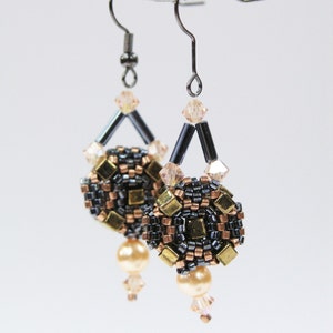Puede incluir: Un par de pendientes colgantes con cuentas negras, doradas y de cobre. Los pendientes tienen un dise&ntilde;o geom&eacute;trico y est&aacute;n adornados con cristales transparentes.