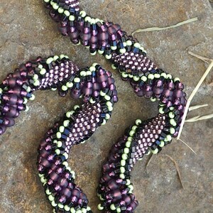 May include: A close-up of a purple and green beaded necklace. The beads are arranged in a spiral pattern and are made of glass. The necklace is handmade and is a unique piece of jewelry.