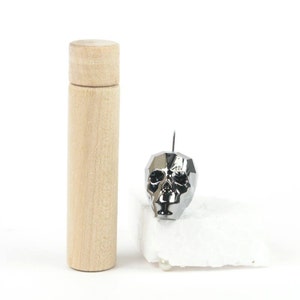 Könnte beinhalten: Eine hellfarbene Holzröhre mit abnehmbarem Deckel steht neben einem silbernen, schädelartigen Objekt. Der Schädel hat ein geometrisches Design und einen kleinen Haken, der sich von oben erstreckt. Der Schädel ruht auf einer weißen, strukturierten Oberfläche.