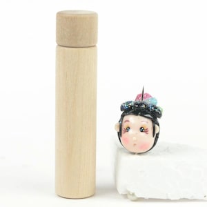 Könnte beinhalten: Eine kleine Holzspule mit runder Oberseite, neben einem handgefertigten Puppenkopf mit schwarzen Haaren, rosafarbenen Wangen und einem bunten Kopfschmuck.