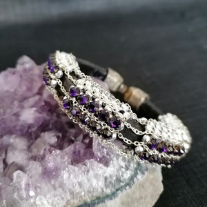 Könnte beinhalten: Ein schwarzes Lederarmband mit silbernen Ketten und violetten Kristallen. Das Armband liegt auf einem violetten Amethystkristall.