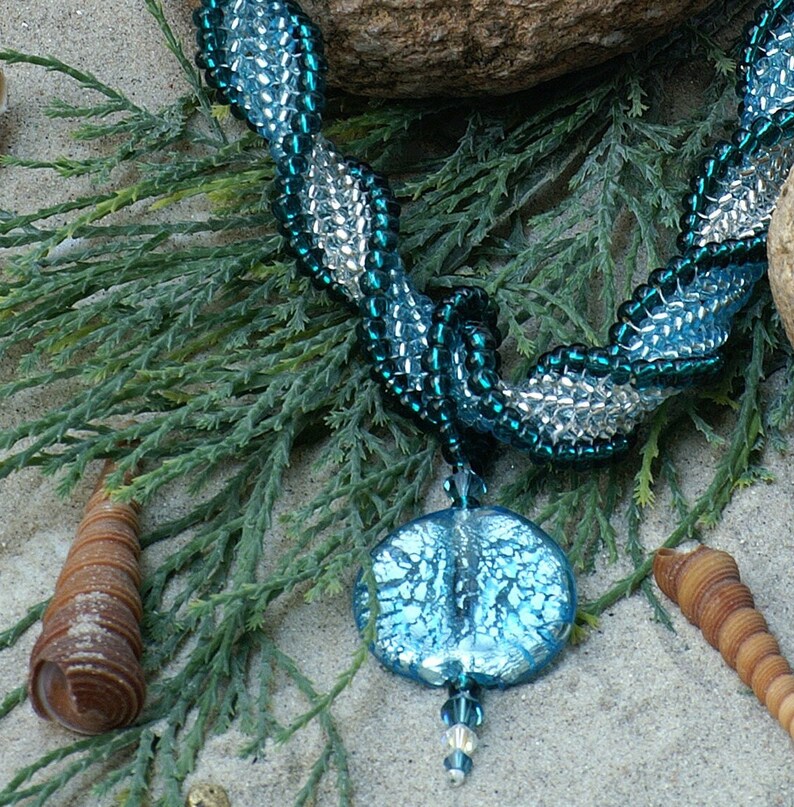 May include: A blue and silver beaded necklace with a large, round, blue glass pendant. The necklace is lying on a bed of green foliage.