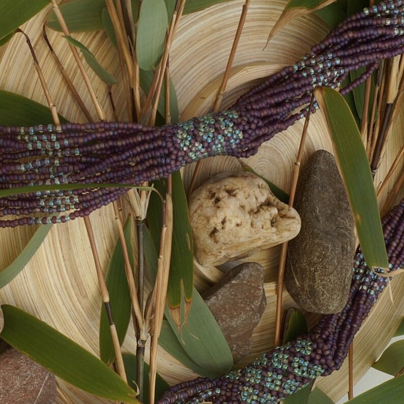 May include: A purple and teal beaded necklace with a woven design. The necklace is displayed on a wooden surface with green leaves.