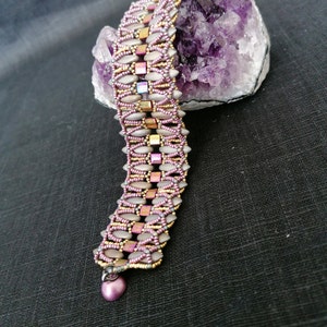 May include: A beaded bracelet with a geometric pattern in shades of pink, gold, and silver. The bracelet is displayed on a purple amethyst crystal.