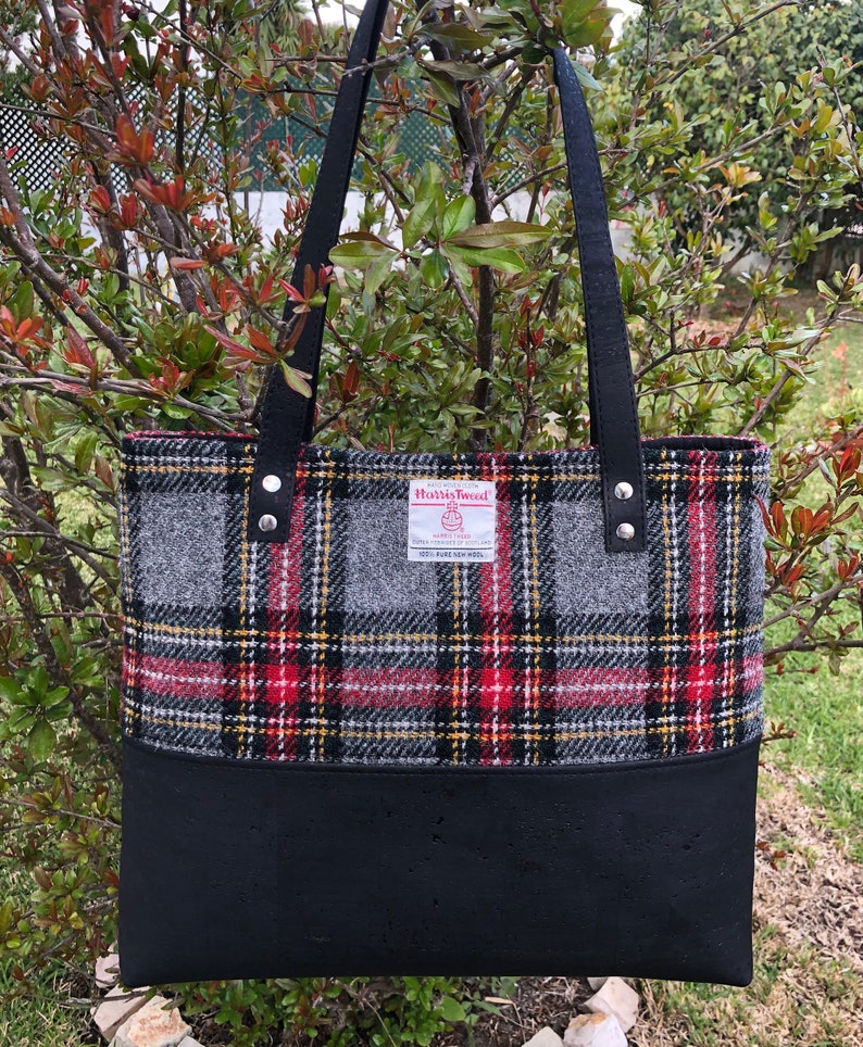 May include: A black tote bag with black leather straps and a red, black, and gray plaid fabric lining. The fabric lining has a label that reads "Harris Tweed" with a logo and the text "HANDWOVEN TWEED 100% PURE NEW WOOL MADE IN SCOTLAND".