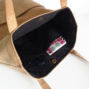 May include: A cork tote bag with a black and white polka dot lining. The bag is open and has a small notebook and glasses inside.