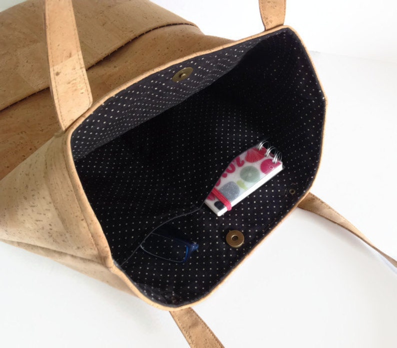 May include: A cork tote bag with a black and white polka dot lining. The bag is open and has a small notebook and glasses inside.