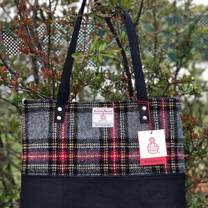 May include: A black tote bag with a black leather bottom and a gray, red, and yellow plaid fabric top. The bag has two black straps and a label that reads "Harris Tweed" with a crest and the words "Handwoven Harris Tweed" and "100% Pure New Wool".