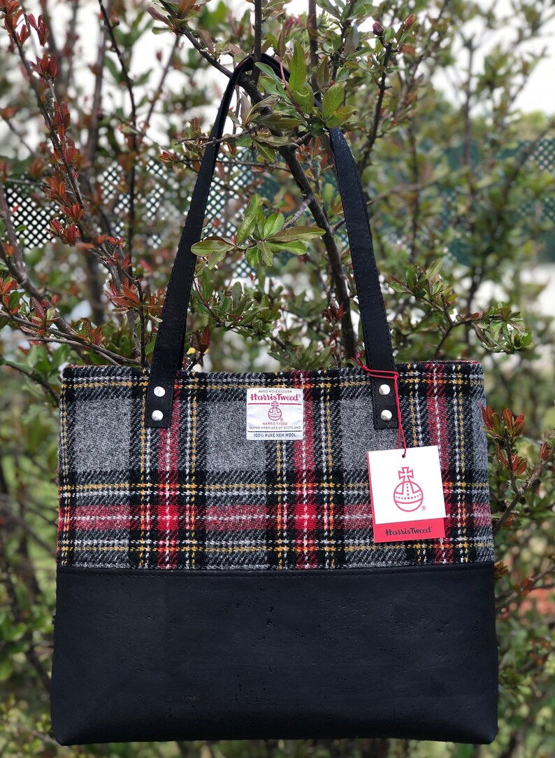 May include: A black tote bag with a black leather bottom and a gray, red, and yellow plaid fabric top. The bag has two black straps and a label that reads "Harris Tweed" with a crest and the words "Handwoven Harris Tweed" and "100% Pure New Wool".