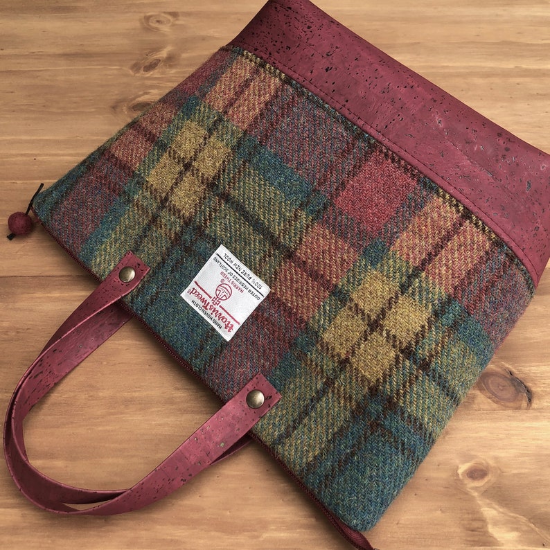 May include: A red and green plaid tote bag with a cork handle and a burgundy cork trim. The bag has a zipper closure and a label that reads "100% Pure New Wool, Made in Scotland, Harris Tweed, Handwoven, Registered Trade Mark".