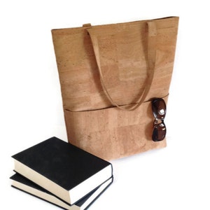 May include: A large cork tote bag with two pockets and a pair of sunglasses tucked into the side pocket. The bag is sitting on a white surface next to two black hardcover books.