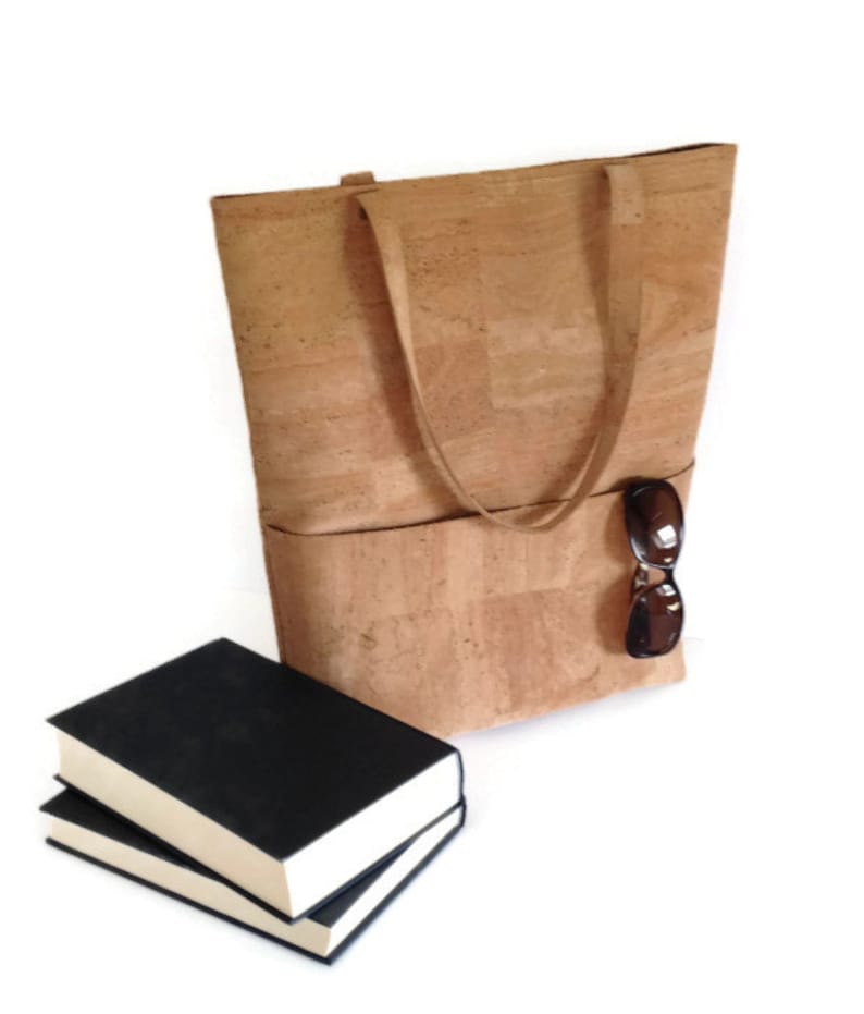 May include: A large cork tote bag with two pockets and a pair of sunglasses tucked into the side pocket. The bag is sitting on a white surface next to two black hardcover books.