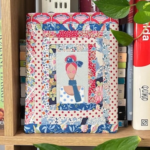 Peut inclure: Une couverture de livre en tissu avec un motif patchwork. Le motif central est une illustration d'une femme aux cheveux roux et une écharpe bleue. Le patchwork comprend des motifs floraux et à pois rouges, bleus et crème.