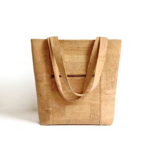 May include: A light brown cork tote bag with two long straps. The bag has a zippered pocket on the front.
