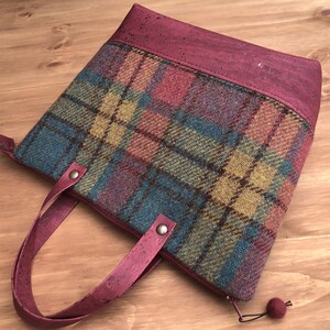 May include: A burgundy tote bag with a plaid pattern and cork handles. The bag has a zipper closure and a small pom-pom on the zipper pull.