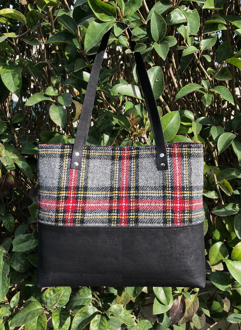 May include: A black tote bag with black leather straps and a plaid pattern in red, yellow, and gray. The bottom half of the bag is black.