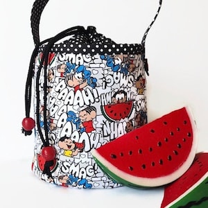 Peut inclure: Un sac à cordon noir et blanc avec un imprimé de personnages et de mots de bande dessinée. Le sac a une poignée noire et des perles rouges. Deux tranches de pastèque sont au premier plan.