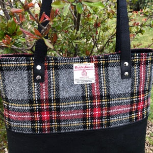 May include: A black tote bag with a red, black, and gray plaid pattern. The bag has a black leather handle and a label that reads "Harris Tweed" with the text "100% Pure New Wool".