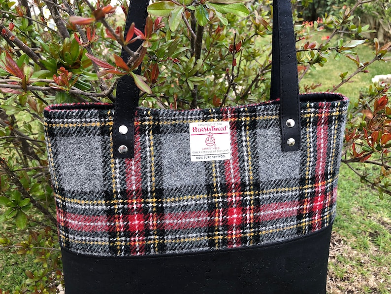 May include: A black tote bag with a red, black, and gray plaid pattern. The bag has a black leather handle and a label that reads "Harris Tweed" with the text "100% Pure New Wool".