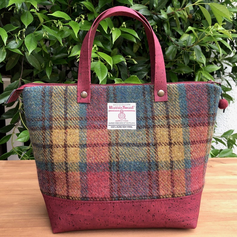 May include: A red, yellow, and blue plaid tote bag with a cork bottom. The bag has a zipper closure and a label that reads "HAND WOVEN ELETH Harris Tweed HARRIS TWEED OUTER HEBRIDES OF SCOTLAND 100% PURE NEW WOOL".