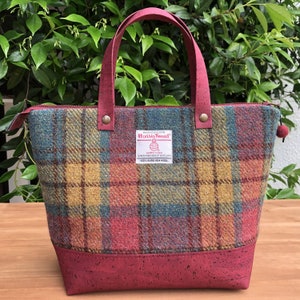 May include: A red, yellow, and blue plaid tote bag with a brown leather handle and a cork bottom. The bag has a zipper closure and a small label that reads "Harris Tweed" with a logo of a stylized sheep.