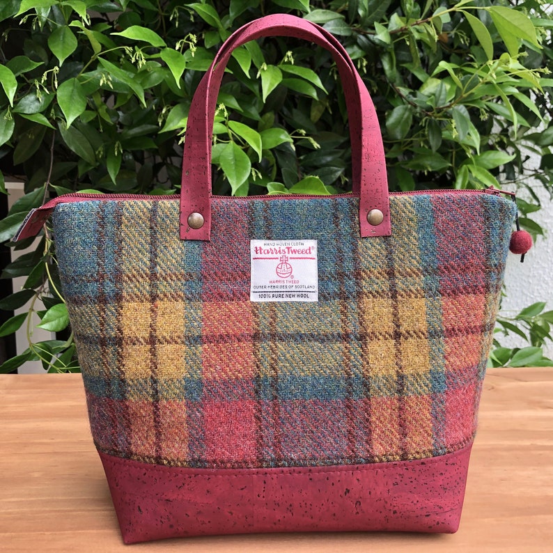 May include: A red, yellow, and blue plaid tote bag with a brown leather handle and a cork bottom. The bag has a zipper closure and a small label that reads "Harris Tweed" with a logo of a stylized sheep.