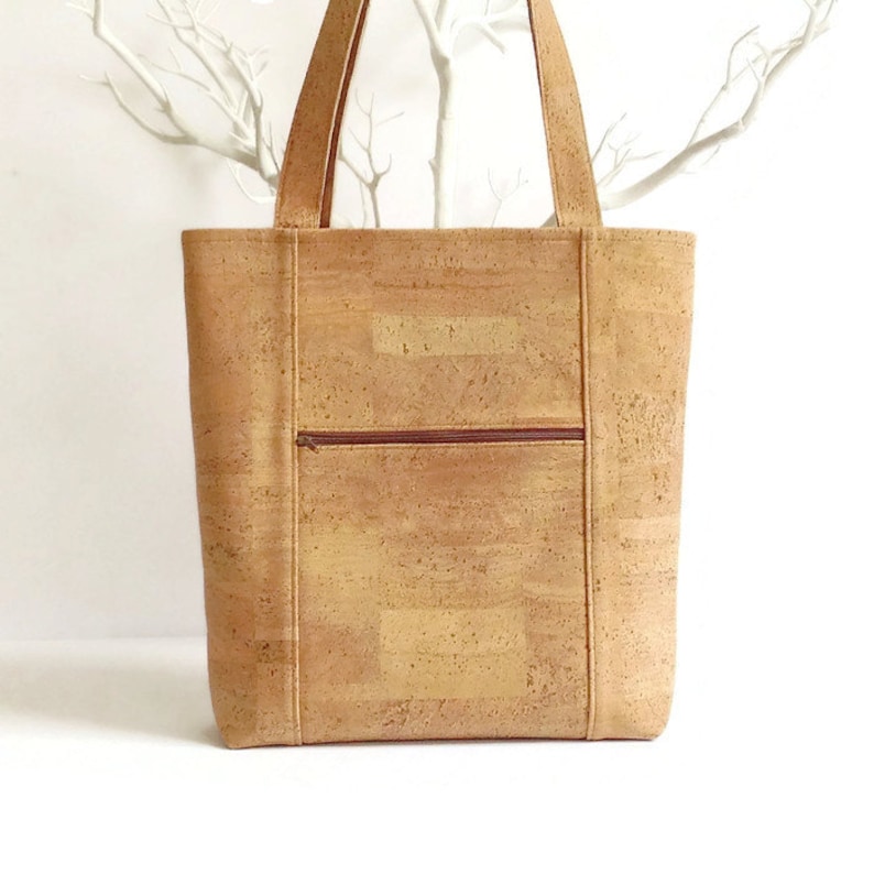 May include: A large, brown cork tote bag with a zippered pocket on the front. The bag has brown stitching and a brown zipper.