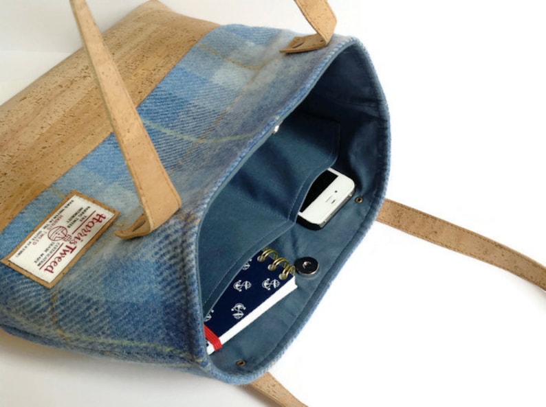 May include: A blue and brown tote bag with a cork handle and a blue tweed fabric body. The bag has a zipper closure and a small pocket on the inside. The bag is open and you can see a phone and a notebook inside.