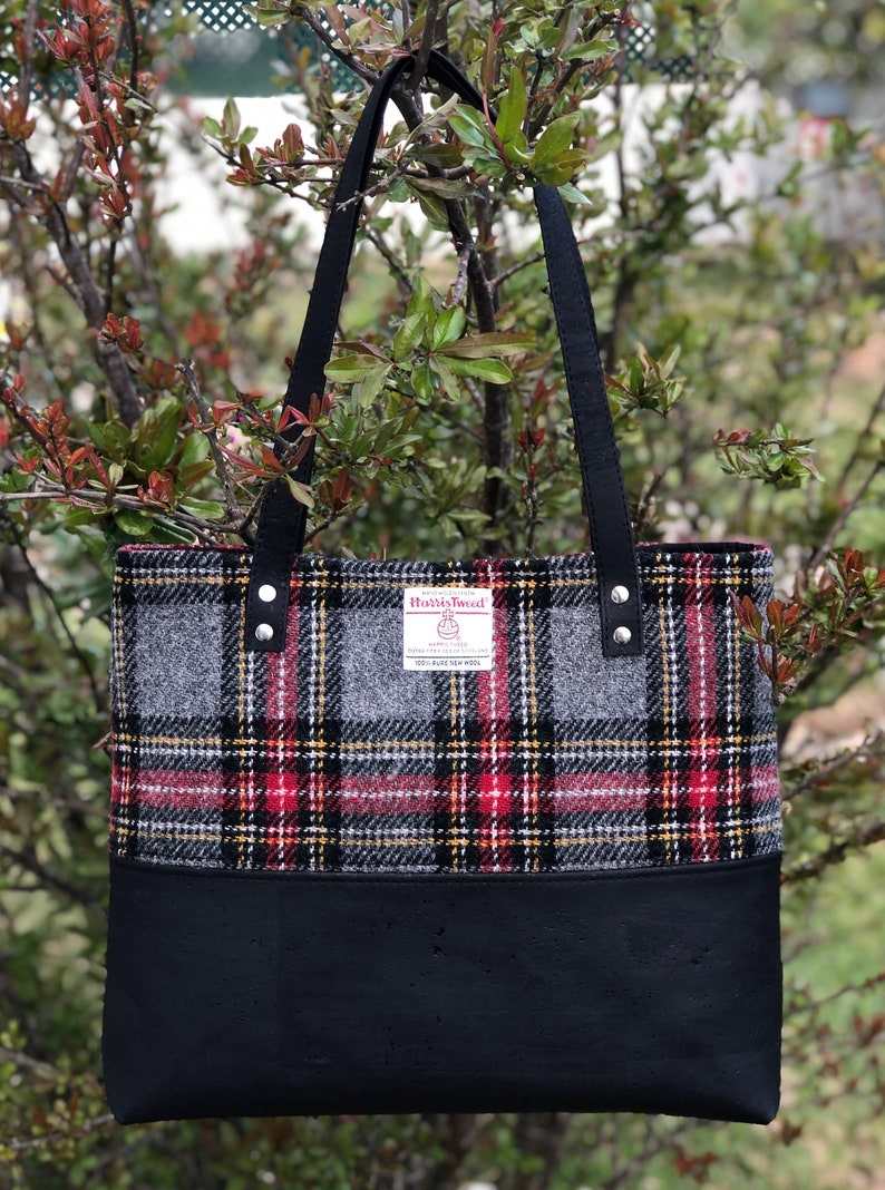 May include: A black tote bag with black straps and a gray and red plaid fabric top. The fabric has a label that reads "Harris Tweed" with the text "100% Pure New Wool" underneath.