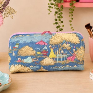Pode incluir: Um estojo retangular acolchoado com fecho de correr, apresentando uma estampa de paisagem azul com ilustrações de pessoas em vestidos amarelos e rosa, um guarda-sol rosa, árvores e mobiliário de exterior. O estojo tem um fecho e rebordo rosa.