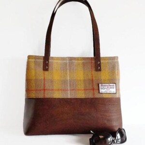 May include: A tote bag with a plaid upper section in yellow, brown, and red, and a brown leather lower section. The bag has brown leather handles and a small label that reads "Harris Tweed". Sunglasses rest at the bottom.