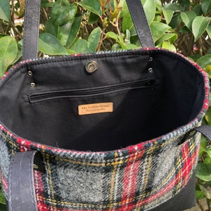May include: Black leather tote bag with a red, green, and gray plaid lining. The bag has a zippered pocket and a snap closure. The label inside reads "My Cotton House Handmade".