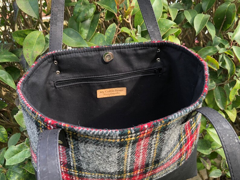 May include: Black leather tote bag with a red, green, and gray plaid lining. The bag has a zippered pocket and a snap closure. The label inside reads "My Cotton House Handmade".